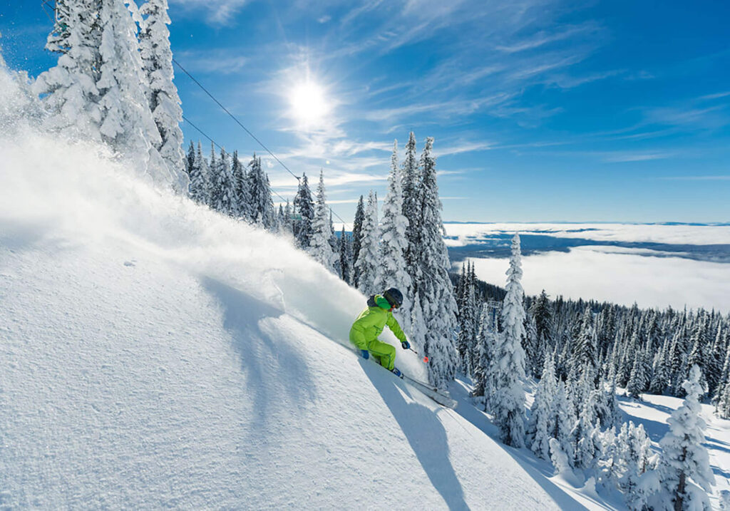 A skilled skier in bright yellow gear skis down a champagne powder snow covered slope with bright blue skies above at Big White Ski Resort.