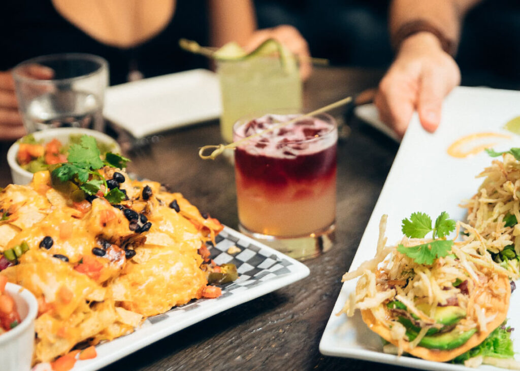 A place of nachos and other appetizers sits with two cocktail drinks.