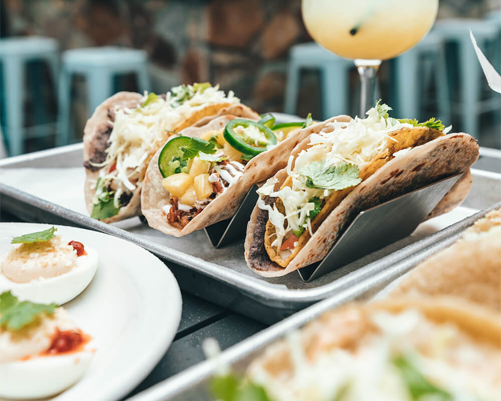 A plate of three fresh tacos sits surrounded by other appetizers and a cocktail drink in the background.