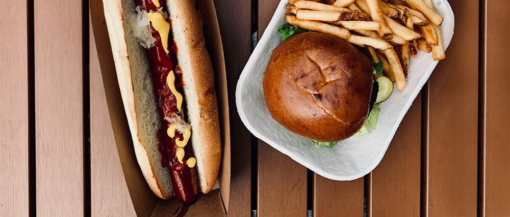 A hot dog with mustard and ketchup and a burger with friends sit on top of a picnic table.