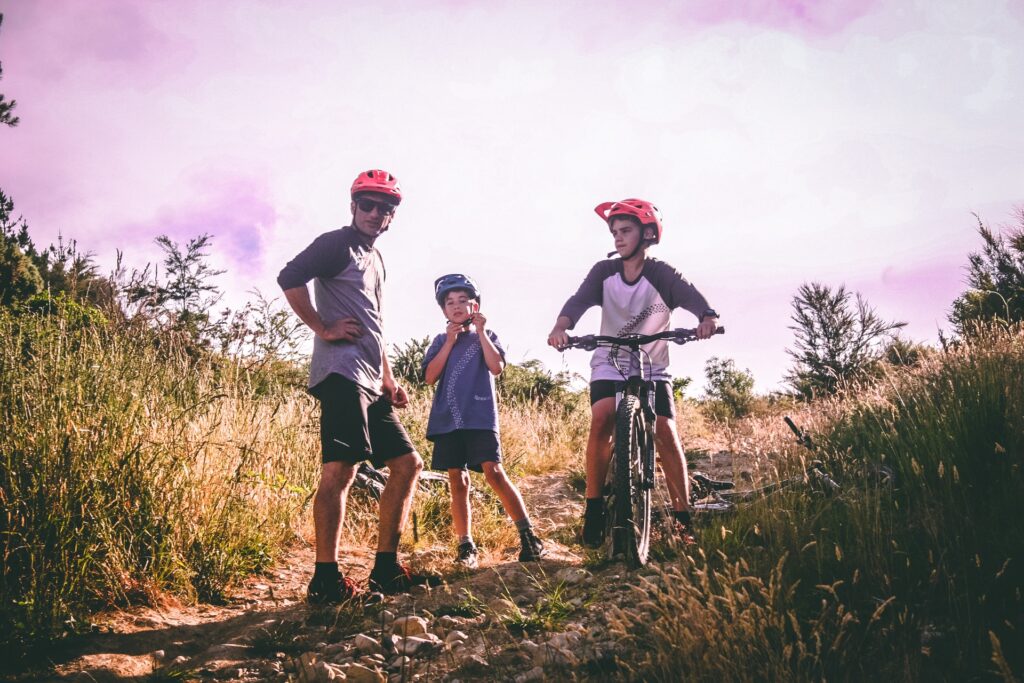 A father and his two young sons stand on a mountain biking trail on a sunny day wearing helmets, while his older son sits on a mountain bike.