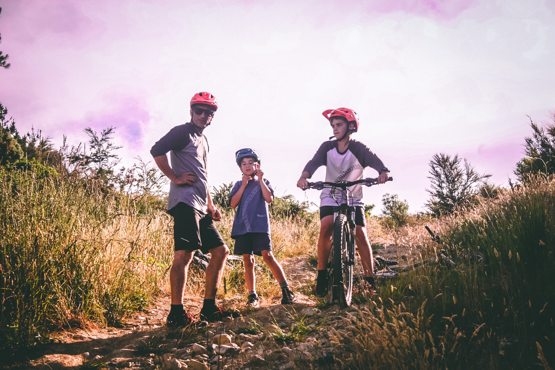 A father and his two kids, one of them on a mountain bike, stand on a dirt trail surrounded by shrubbery on the top of a mountain while on a family vacation.