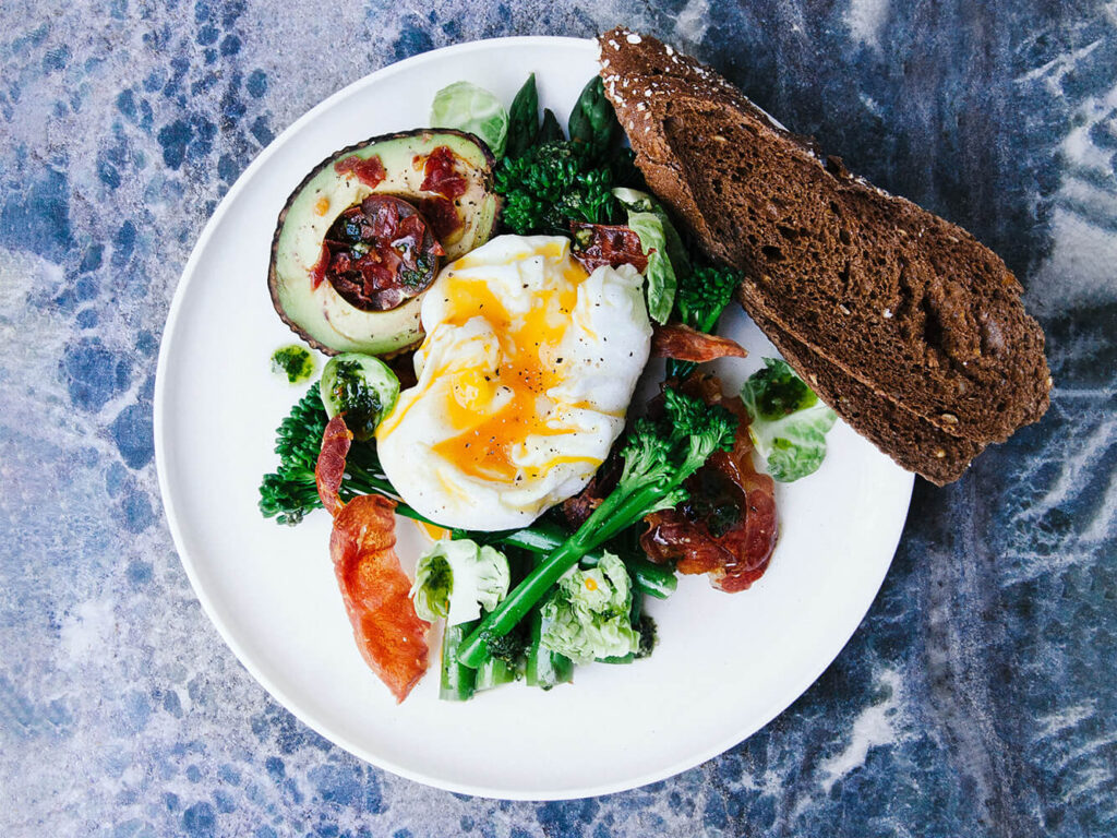 A plated dish with a roasted salad, avocado, poached egg, and piece of toast sits on a blue marbled table.