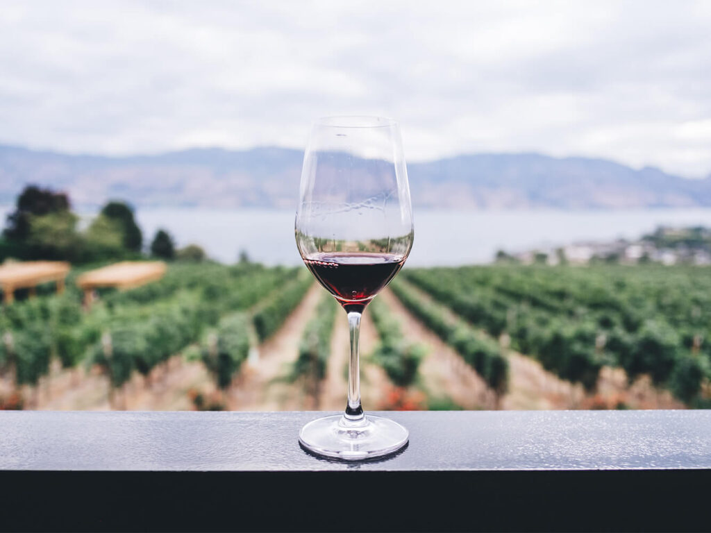 A glass of red wine sites on a railing overlooking a beautiful wine vineyard and the Okanagan lake in BC. During a private sommelier wine tasting, you'll be able to enjoy many of the Okanagan's best wines, and others, during your stay with Luxury Mountain Vacation Rentals at Big White Ski Resort.
