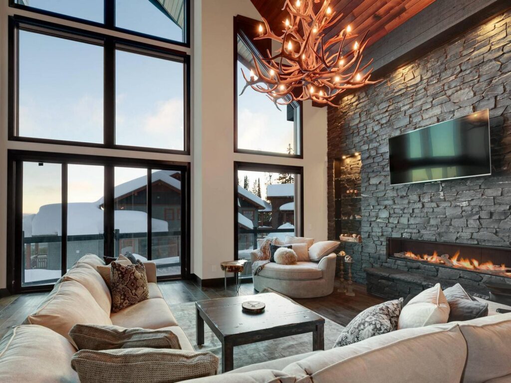 Large open modern ski chalet living room with an antler chandelier, stone fireplace and floor to ceiling windows.