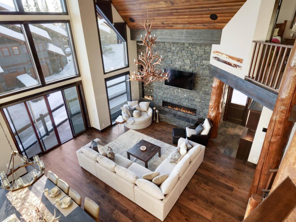 Overview of the great room/living room with an antler style chandelier, cozy couch and fireplace and large windows, with a luxury ski chalet feel, a vacation rental managed by Luxury Mountain Vacation Rentals.