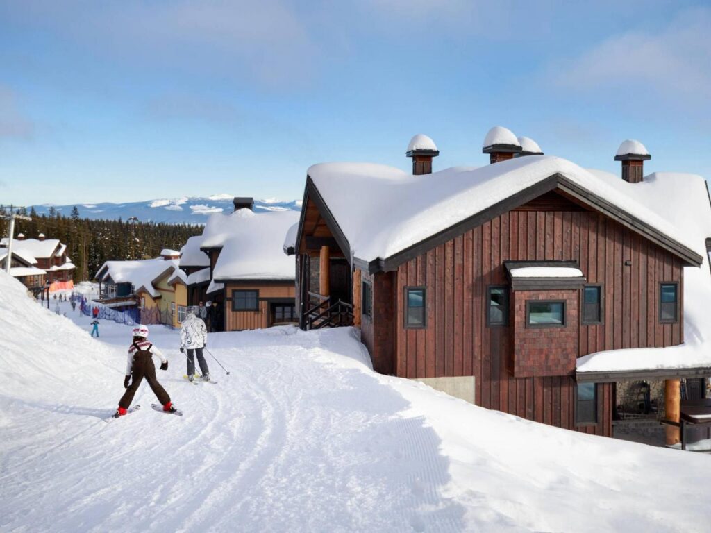 A family of skiiers ski along a snowy path past some luxury ski chalet rentals at Big White Ski Resort on a sunny bluebird day.