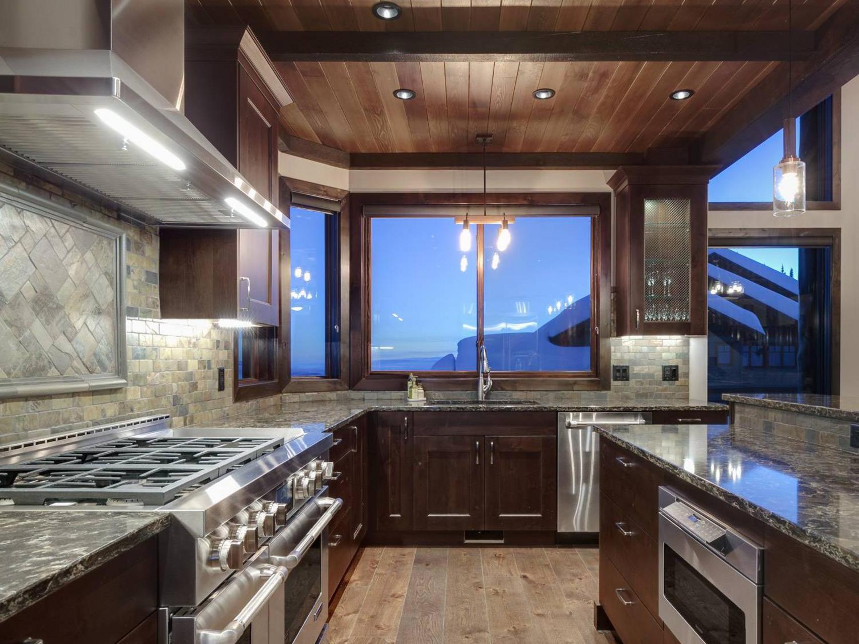 A large, fully-equipped gourmet kitchen with dark wooden cabinets, grey marble countertops, high-end appliances and large windows in a vacation rental by Luxury Mountain Vacation Rentals at Big White Ski Resort.
