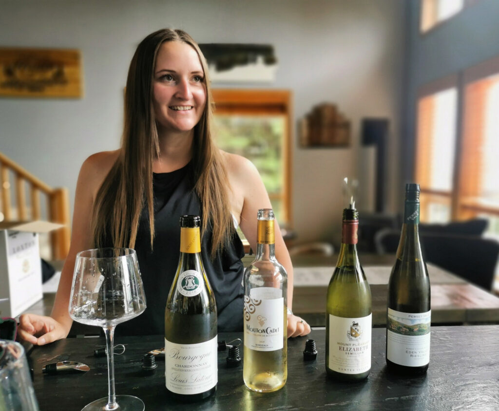 Maddie, a private sommelier at Big White Ski Resort, wears a black shirt and stands with a set of bottles of wine on a counter, preparing a private wine tasting for guests of Luxury Mountain Vacation Rentals.