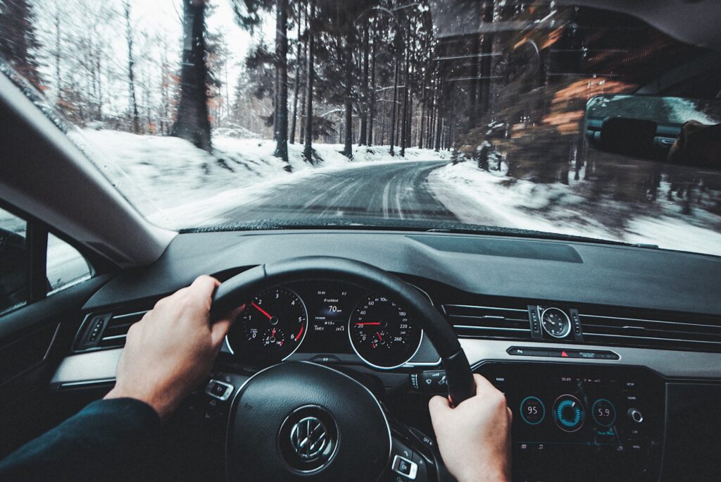 A man holds a steering wheel as he drives a car along the road with snow and trees on either side.