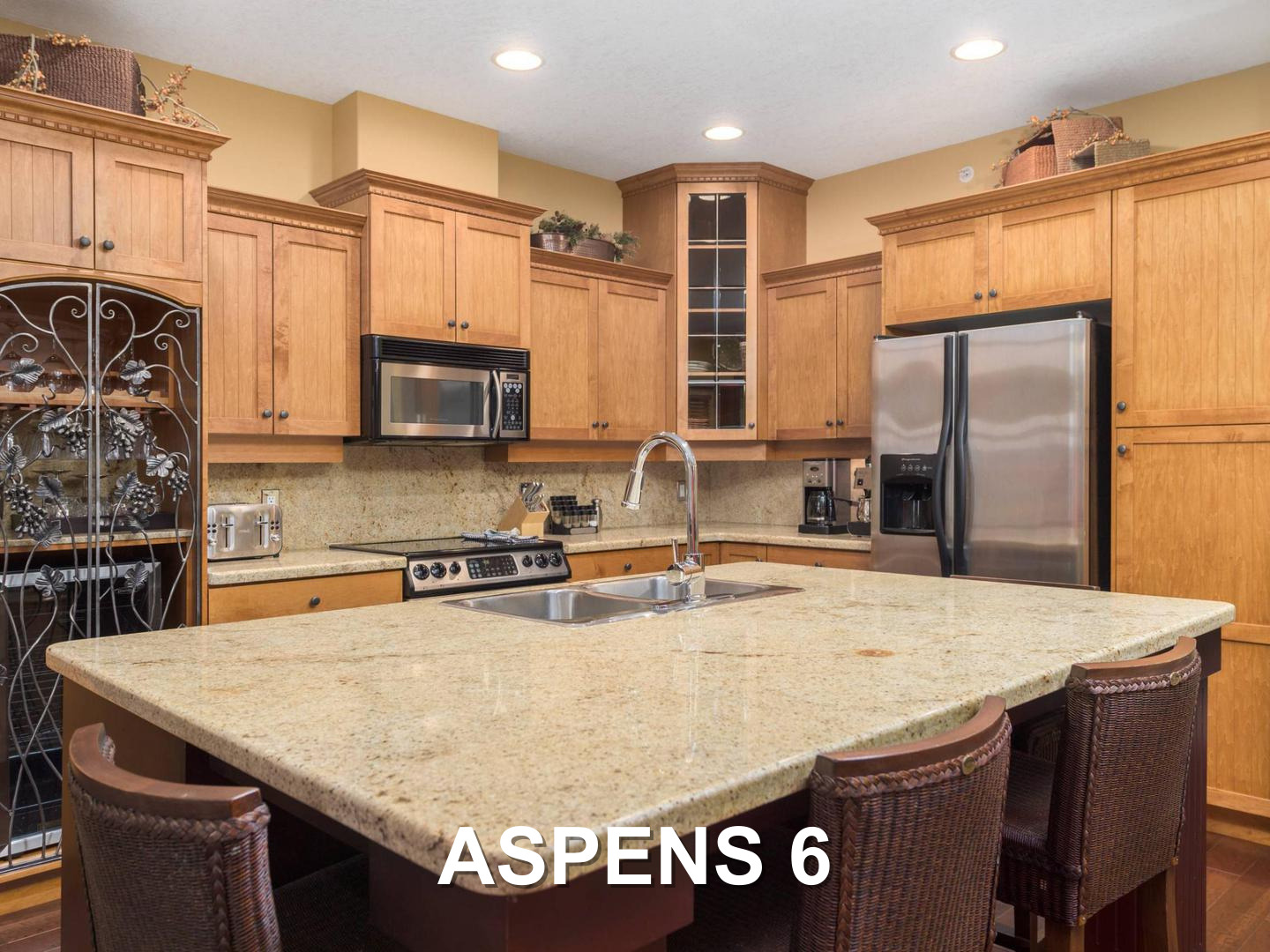 The Aspens 6 luxury, gourmet kitchen, with a stainless steel fridge and oven, a microwave, and a large granite topped kitchen island, with warm wooden cabinets, located at Big White Ski Resort and managed by Luxury Mountain Vacation Rentals.