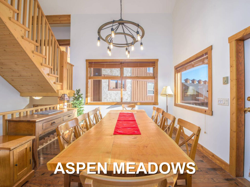 Aspen Meadows' dining room, with a large, long dining table of warm pine wood, a modern chandelier, bright natural light and a wooden staircase on the left leading to the second floor of the vacation rentals, located at Big White Ski Resort and managed by Luxury Mountain Vacation Rentals.