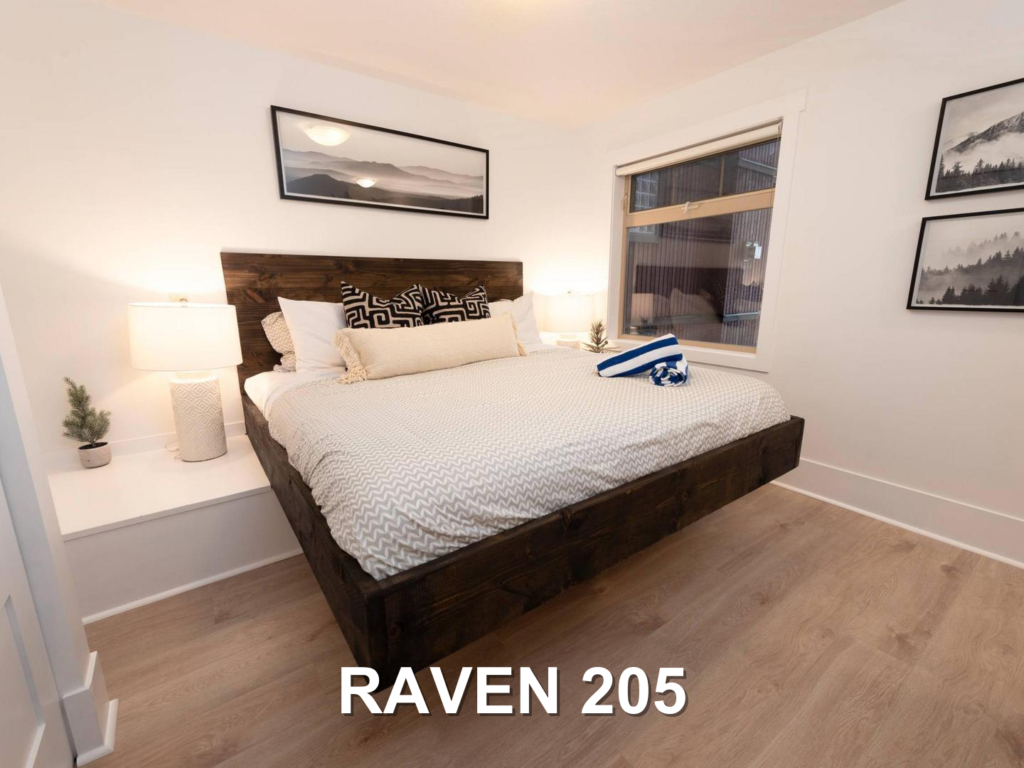 One of the Raven 205's bedrooms, with a raised bed on a dark wooden bedframe, luxury white linens, bright lighting and white walls and a comfortable space, located at Big White Ski Resort and managed by Luxury Mountain Vacation Rentals.