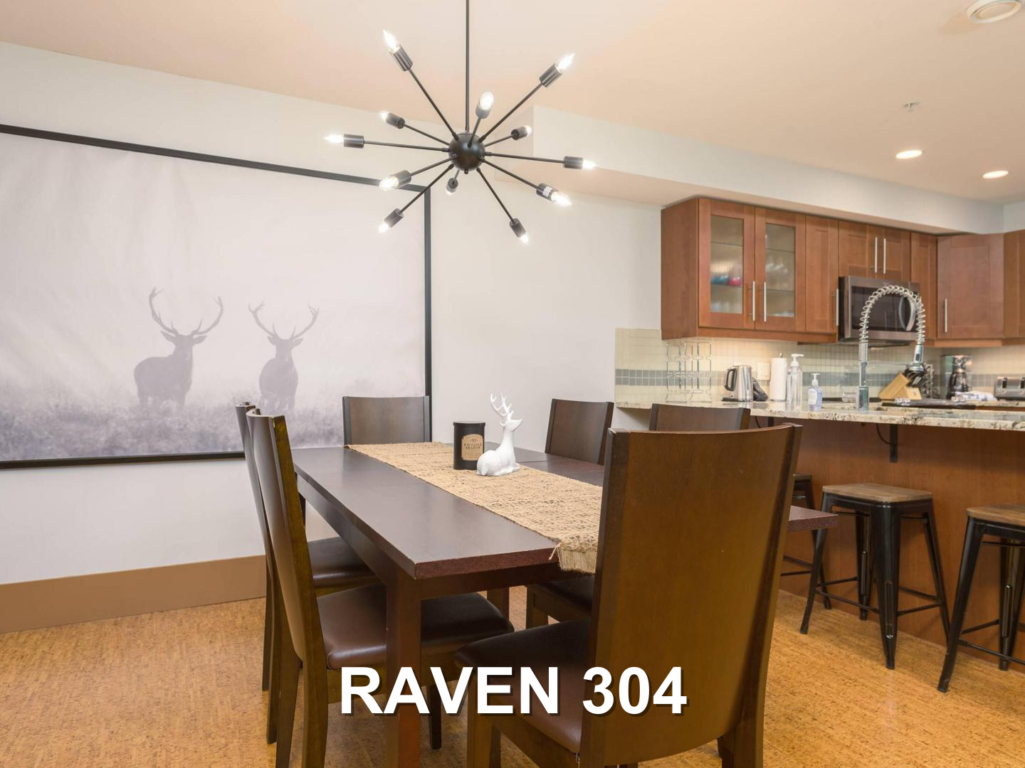 The Raven 304 dining room, with a dark wooden table and modern chandelier above, and the kitchen in the background, located at Big White Ski Resort and managed by Luxury Mountain Vacation Rentals.