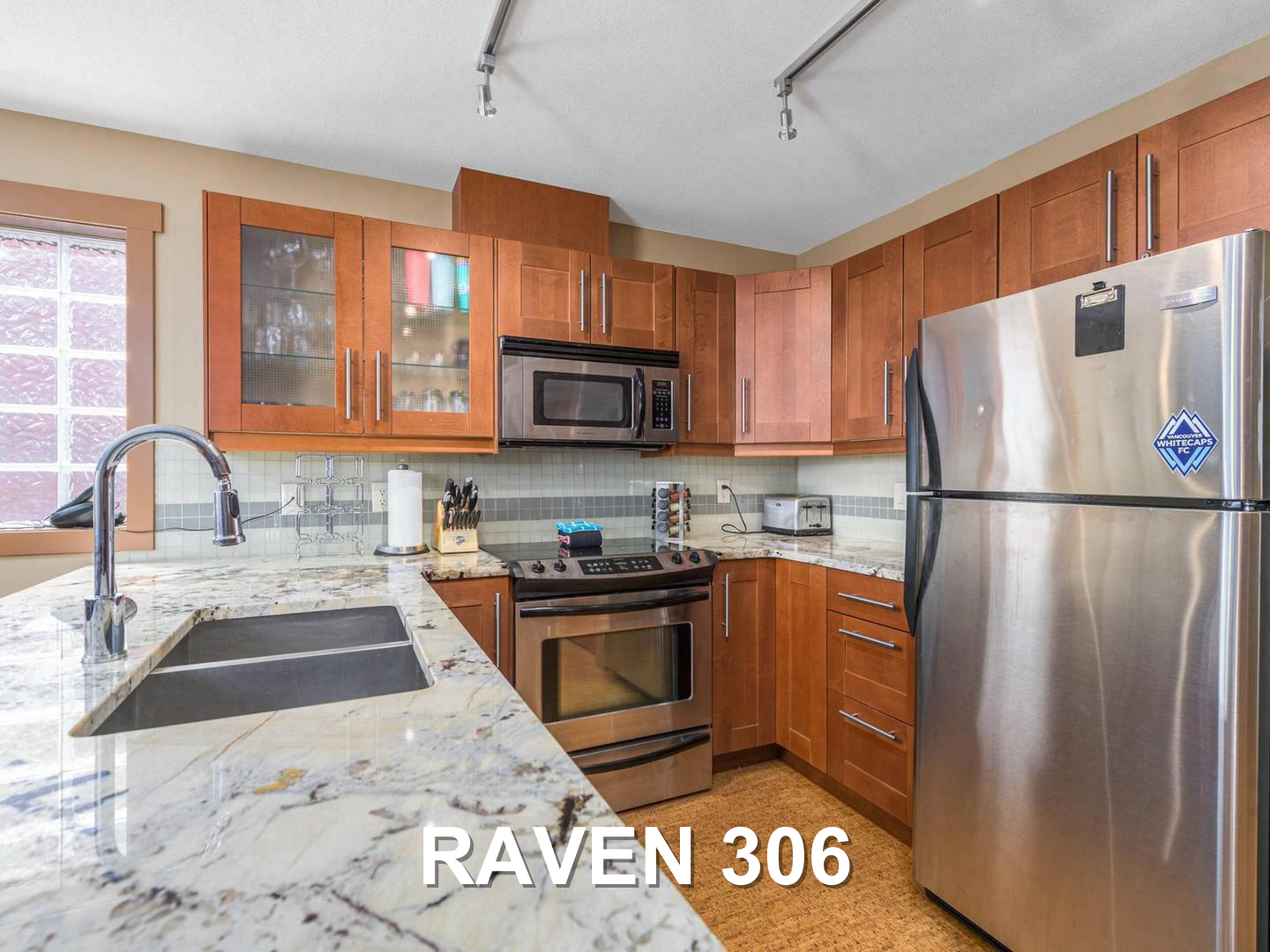 The Raven 306 gourmet kitchen, which is fully equipped with stainless steel appliances, and has wooden cabinets and white marble countertops, located at Big White Ski Resort and managed by Luxury Mountain Vacation Rentals.