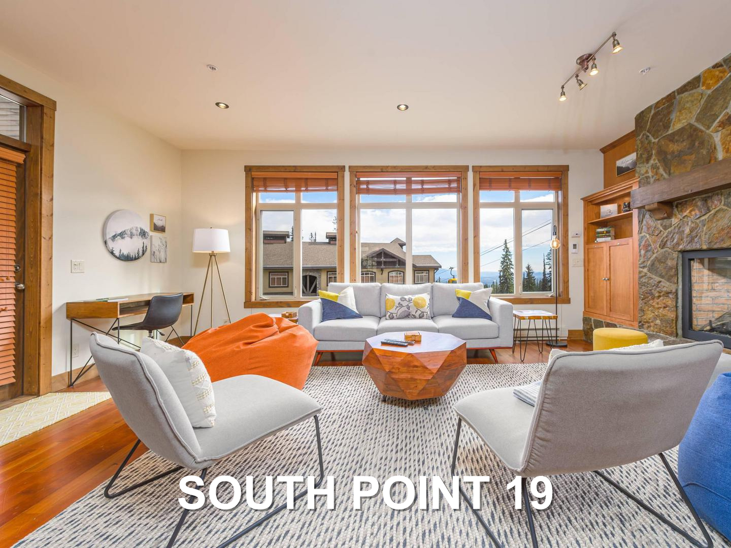 South Point 19's luxury living room, with a stone fireplace, modern grey chairs and couches, and large window with natural light, located at Big White Ski Resort and managed by Luxury Mountain Vacation Rentals.
