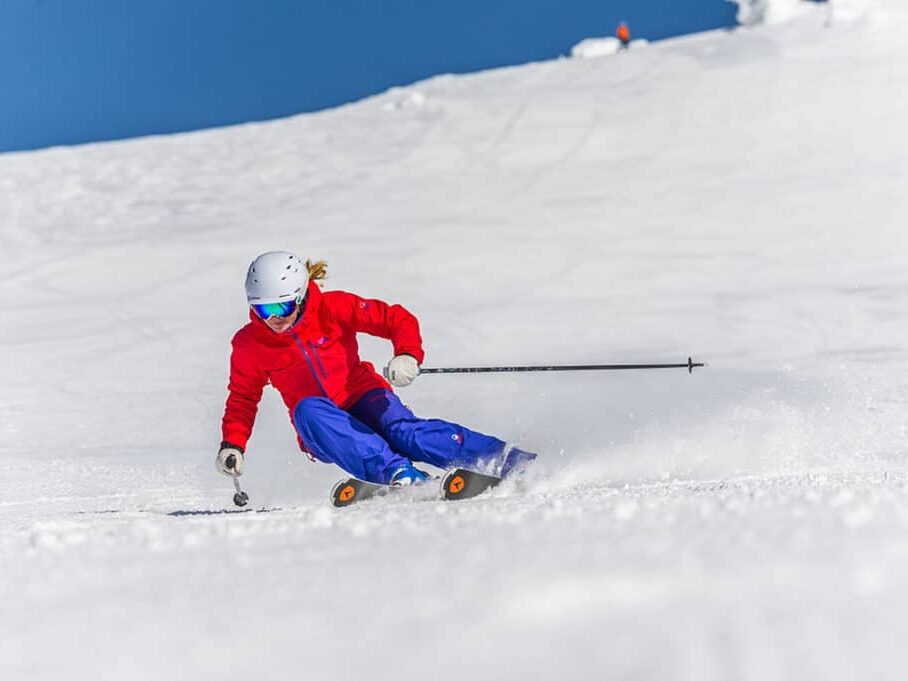A skier dressed in red and blue skis down a snowy trail on the side of the mountain at Big White Ski Resort, with bright blue skies and freshly packed, champagne powder snow.