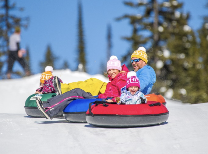 A family of four in winter gear are smiling and laughing as they go tubing at Big White's Tube Park.