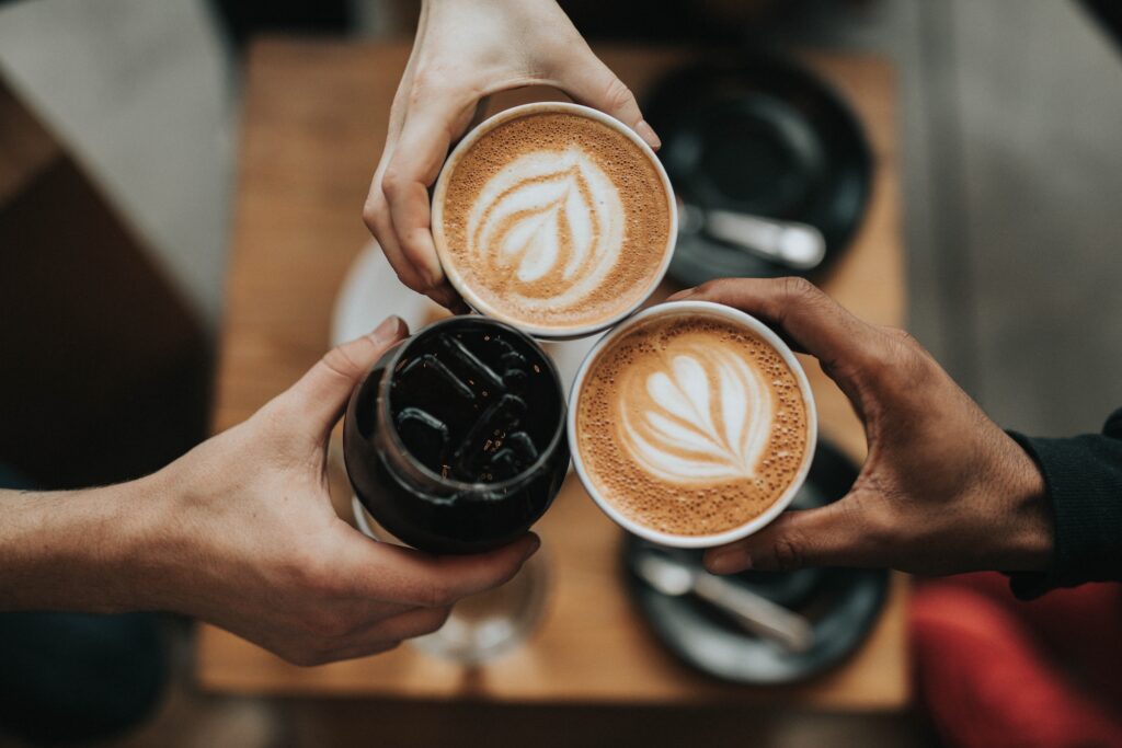 An overhead view of three people cheersing their coffee drinks with two lattes and an iced coffee over a table.