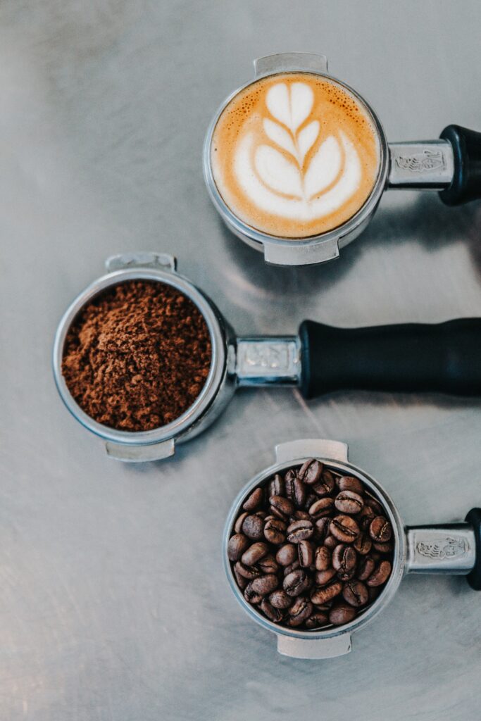 Three espresso porta filters sit on a metal table, one filled with unground cofee beans, one with ground espresso, and another with latte art.