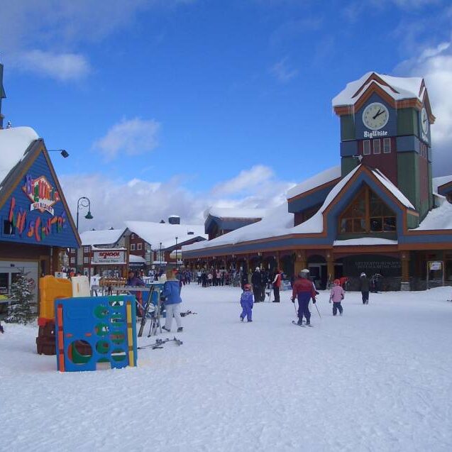 Big White Ski Resort's Village Centre on a suny day with fluffy snow on the crowd and skiers and snowboarders walking around the village, with Big White's big clock in the background and the Kids Centre on the left.