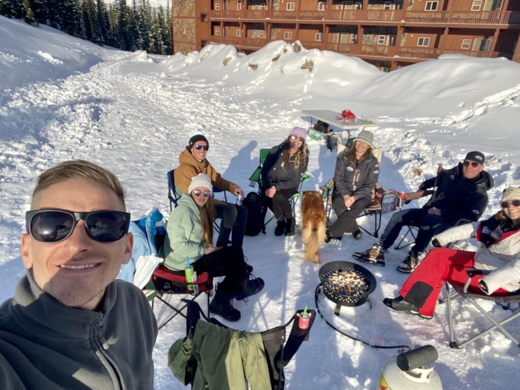 A staff group photo of the staff members at LMVR Luxury Mountain Vacation Rentals, posing outside on a snowy day around a campfire at Big White Ski Resort.