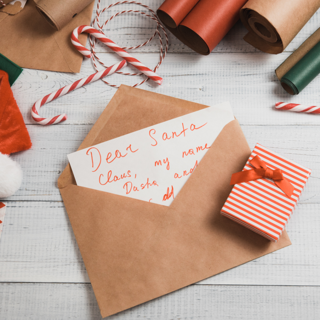 A letter to Santa with the words "Dear Santa" in red ink, placed slightly inside a brown envelope on top of a white wooden table with Christmas wrapping paper, decorations and candy canes around it.