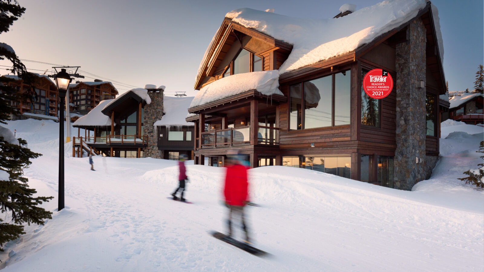 Moonshine Chalet's exterior at sunset on a snowy day with skiiers and snowboarders going by, a luxury mountain chalet rental managed by Luxury Mountain Vacation Rentals at Big White Ski Resort.
