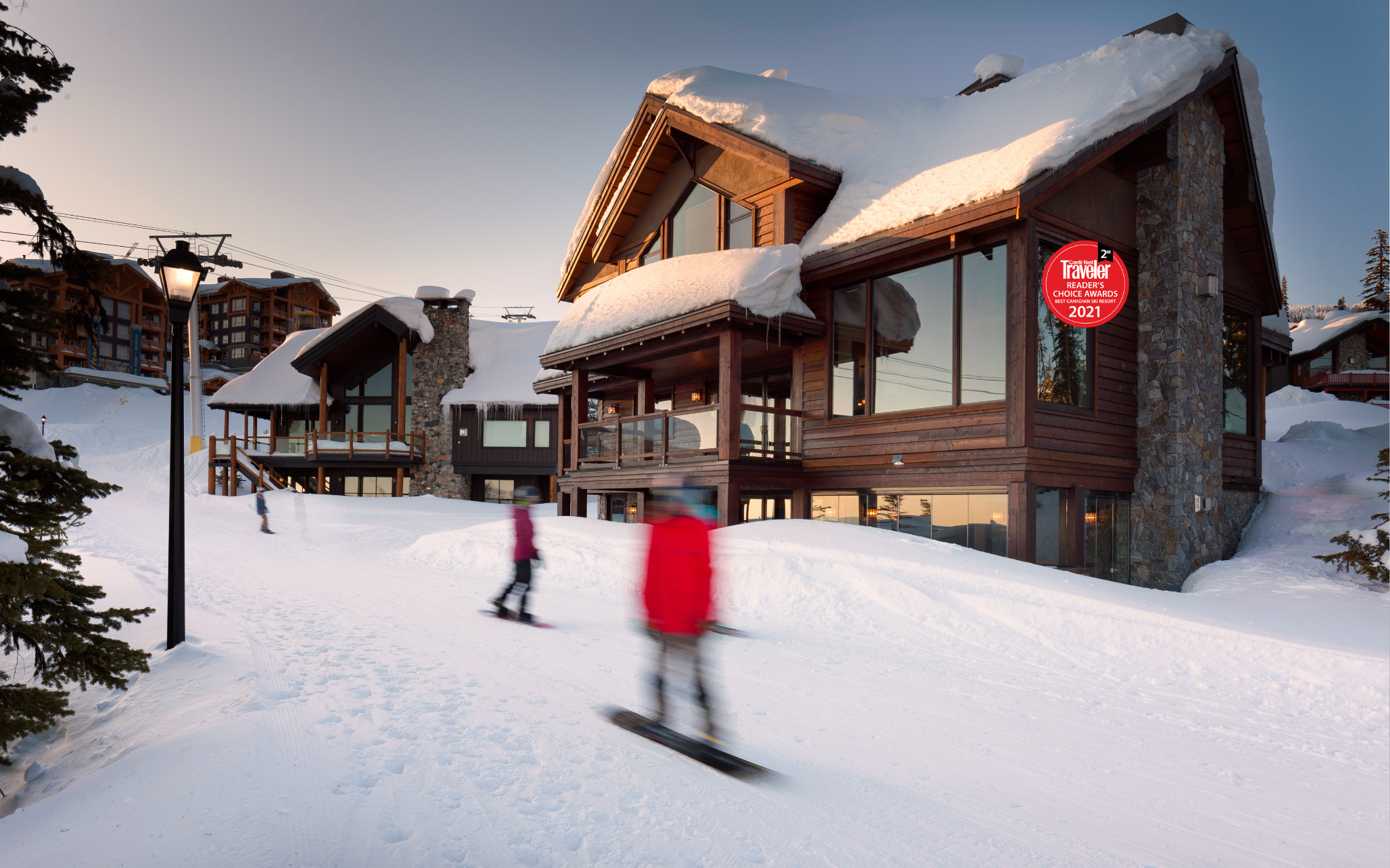 Moonshine Chalet's exterior at sunset on a snowy day with skiiers and snowboarders going by, a luxury mountain chalet rental managed by Luxury Mountain Vacation Rentals at Big White Ski Resort.