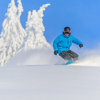 A snowboarder in a blue jacket snowboards down a champagne powder, snowy slope with snow-covered trees in the background and bright blue skies above while at Big White Ski Resort, perfect conditions for snowboarding or skiing in January.