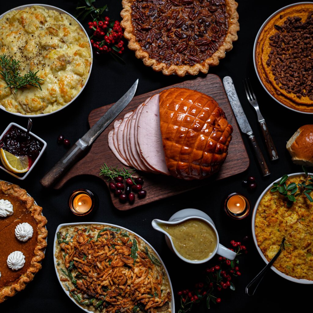 A Christmas Dinner feast with roasted ham, roasted vegetables, mashed potatoes, gravy, and delicious pumpkin pie laid out on a wooden table.