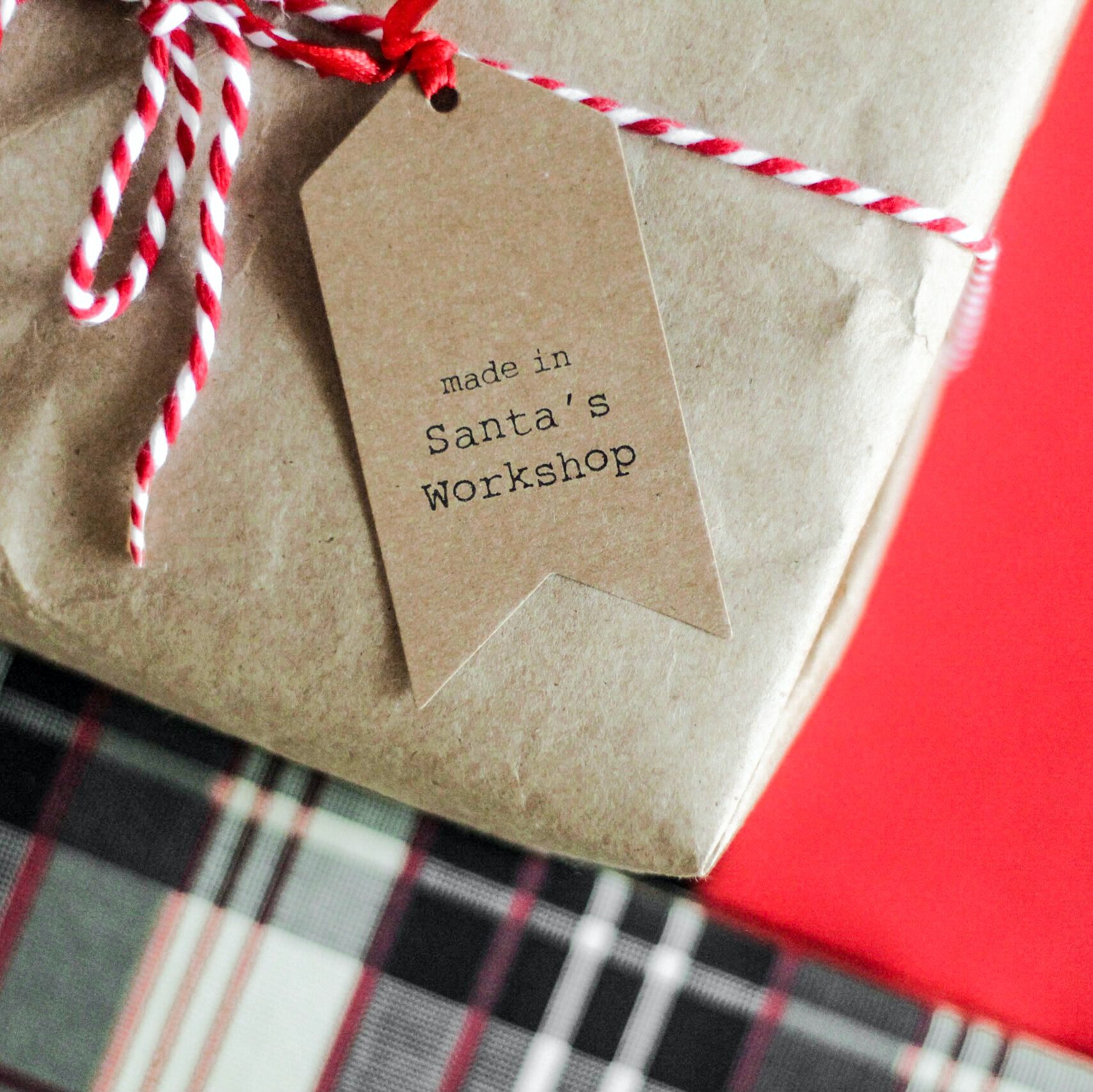 A Christmas gift wrapped in brown paper wrapping and a red and white striped string with a gift label that reads "made in Santa's workshop" on top of a red table.