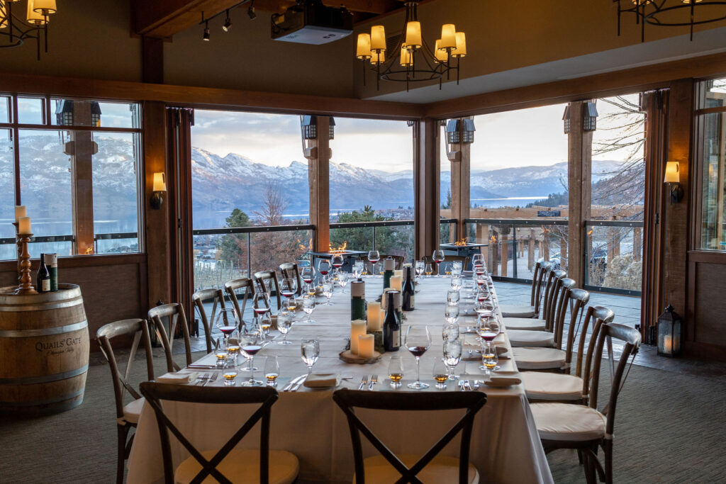A beautiful dining room with a large 25 person table set with wine glasses and fancy dishes on a white table cloth with warm ambient lighting and beautiful lake and vineyard views in the background in winter at the Old Vines Restaurant at Quails' Gate Winery.