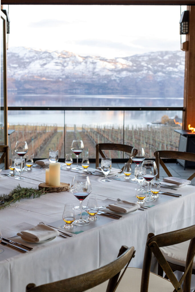 A fancy dining table with a white table cloth, candles and wine glasses is set up in the Old Vines Restaurant dining room with floor to ceiling windows featuring beautiful views of the Quails Gate Winery vineyards and Okanagan lake in winter.