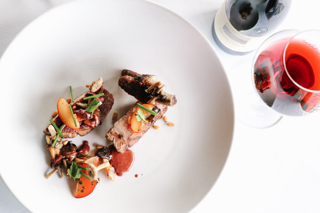 A gourmet dish of vibrant coloured roasted veggies and seared meat on a white plate with a wine bottle and glass of red wine next to it.