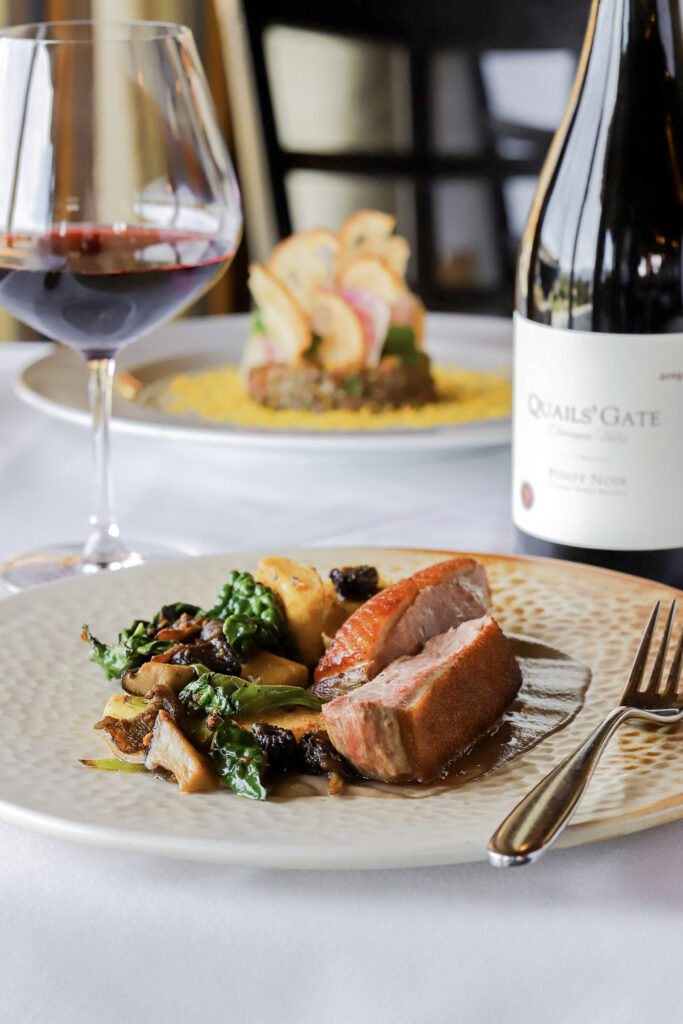 A gourmet dish of seared salmon and vegetables arranged on a ceramic plate wth a glass of red wine and bottle of wine in the background.